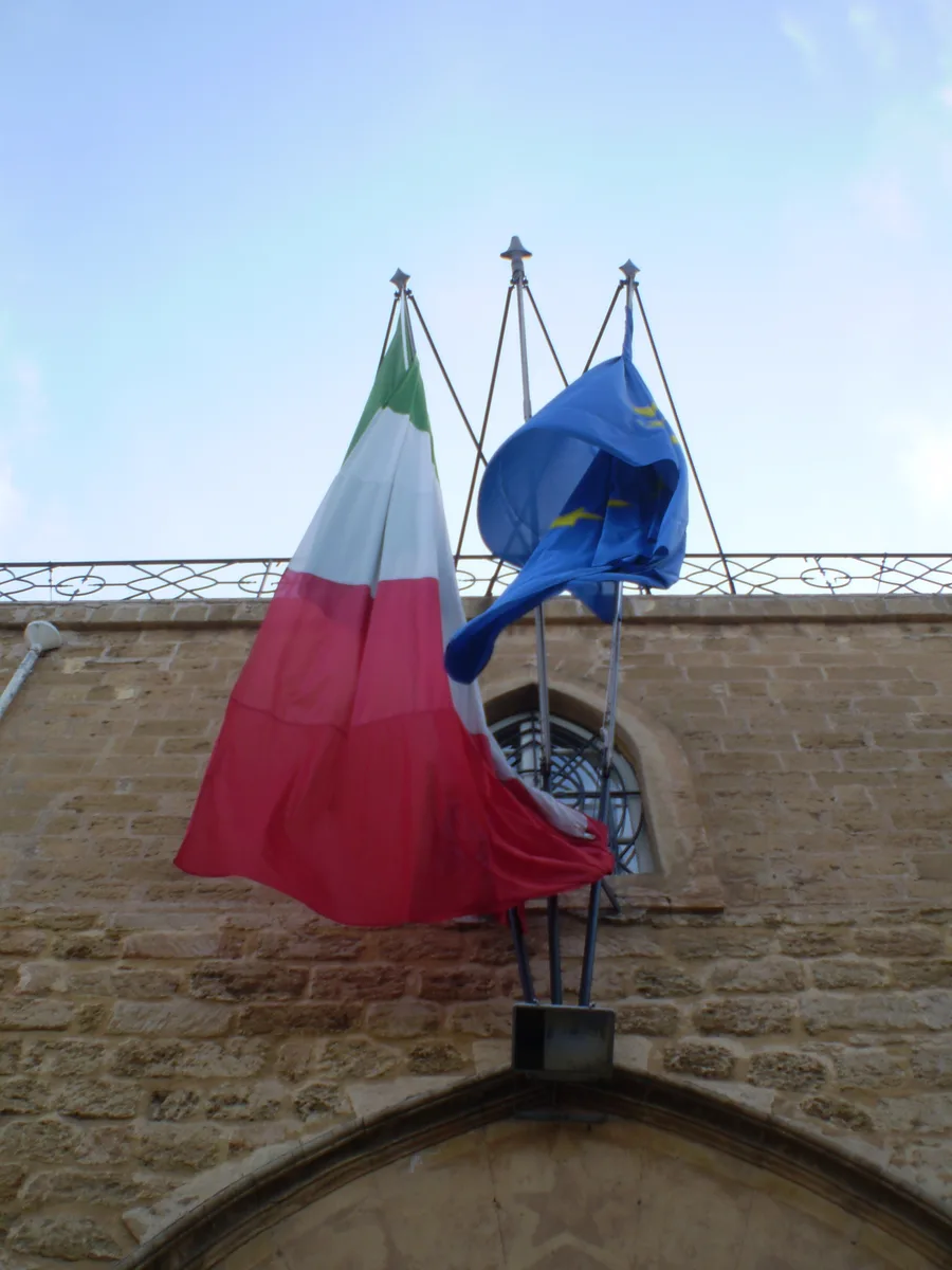 La bandiera dell'Unione Europea, con dodici stelle dorate su sfondo blu, simboleggia unità, solidarietà e armonia tra i popoli d'Europa. Il numero di stelle è fisso a dodici, rappresentando la perfezione e la completezza, piuttosto che il numero degli stati membri. Questa bandiera è esposta in modo prominente presso istituzioni come la Commissione europea, che svolge un ruolo cruciale nel sostenere il piano di ripresa dell'Italia. (Andrea Pavanello, CC BY-SA 3.0 it)